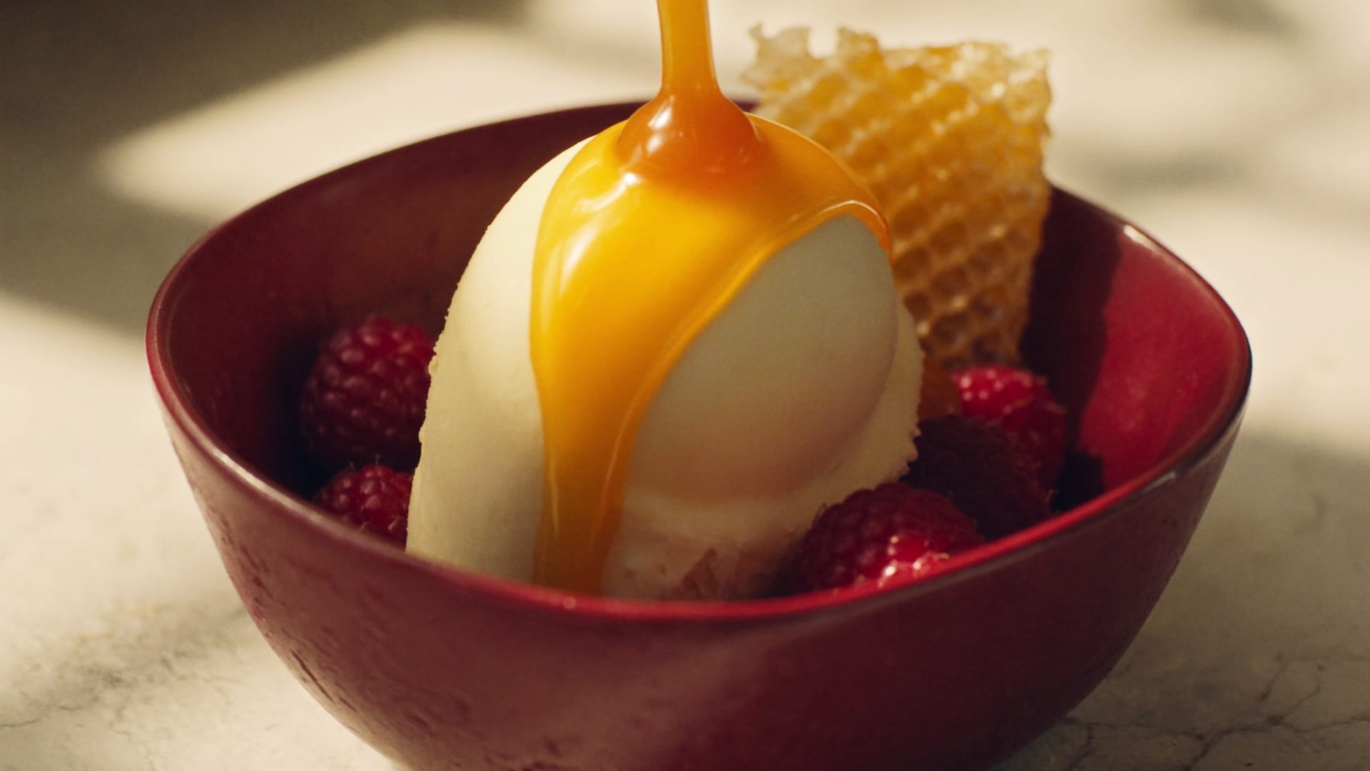 Caramel pouring over a scoop of gelato in a burgundy bowl, with raspberries and honeycomb